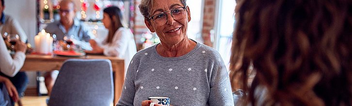 Frau mit Kaffeetasse in der Hand im Gespräch, im Hintergrund Menschen an einem Tisch